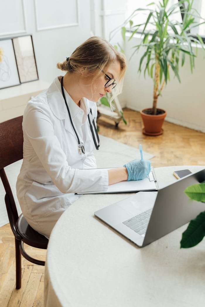 A female doctor in a white coat writes notes at a desk with a laptop.