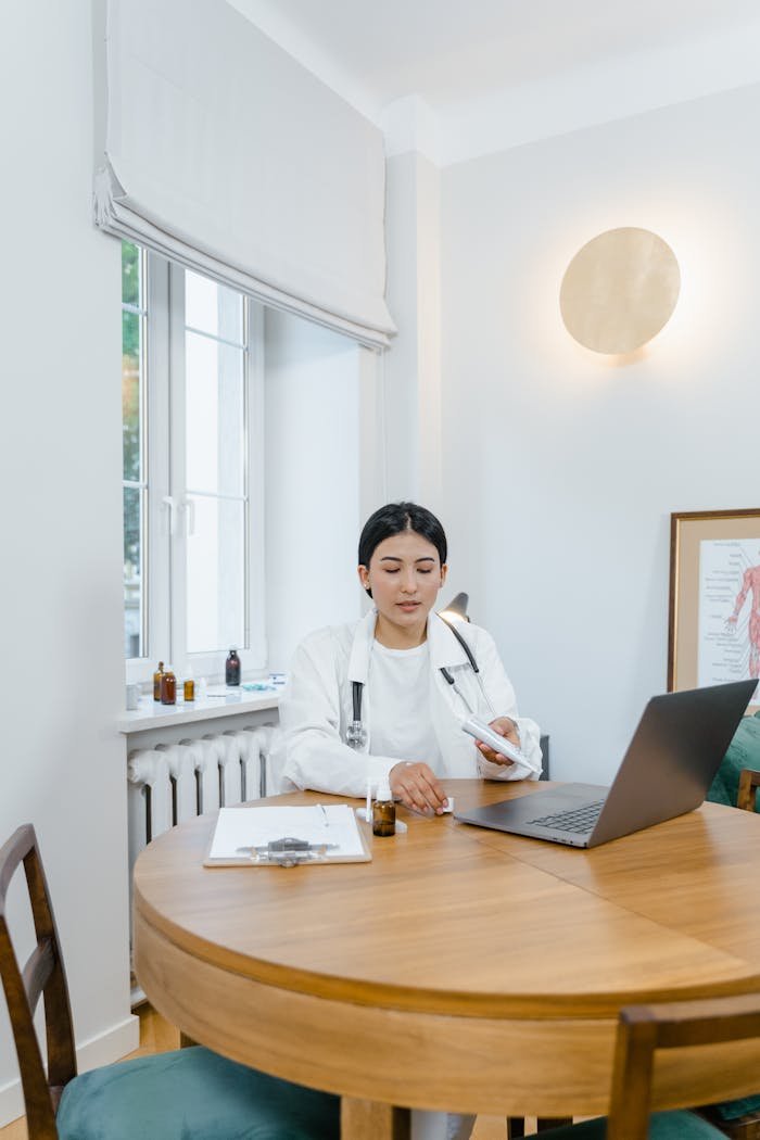 A female doctor in a white coat conducts an online consultation using a laptop in a modern home office.